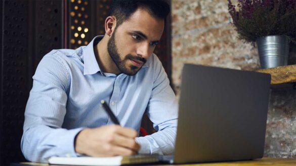 A man writing notes while looking at a laptop