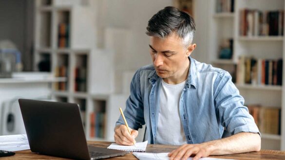 A man looking at a laptop while writing on a notepad