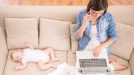 Maternity redundancy changes: mother with laptop while baby sleeps beside her on a sofa.