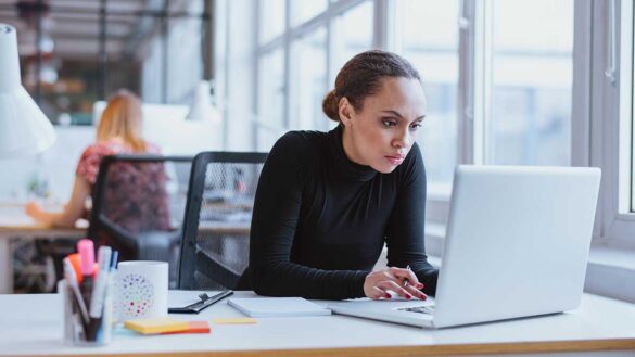 A woman concentrating at a laptop in an open plan office, with a notepad and pen pot beside her on the table
