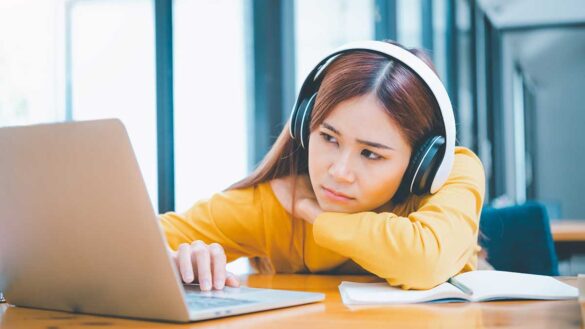 A young woman wearning headphones looking board and tired while working on a laptop