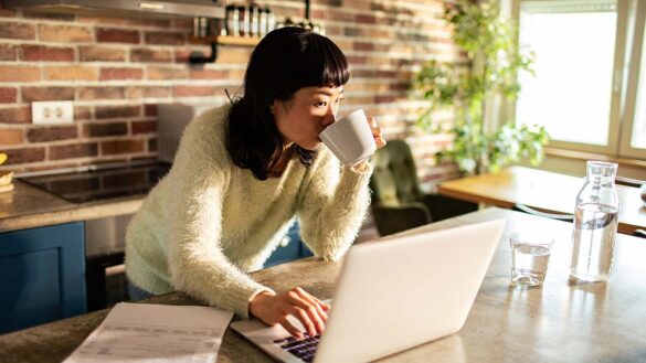 A woman drinking from a mug while working from her kitchen.. A laptop is open on the table