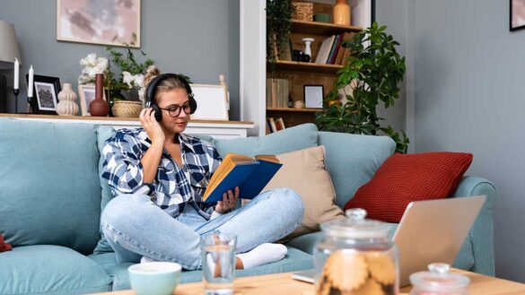 Four-day week skills development: A woman sitting on a sofa. She has headphones on and is reading a book, with a laptop just out of reach