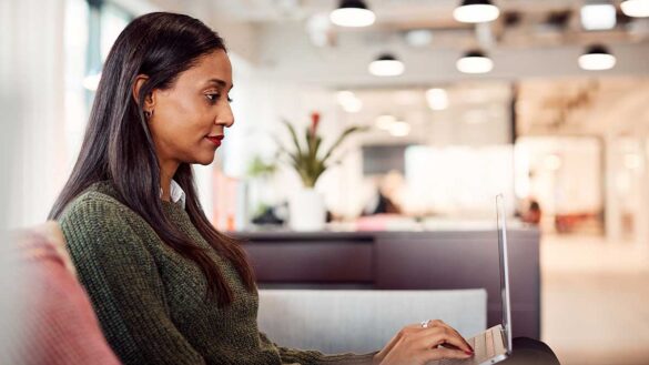 A woman using a laptop while sitting on a sofa in an open plan office