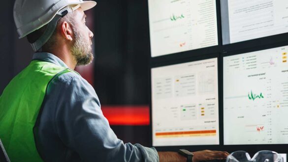 A man in a hard hat and hi-vis jacket sitting at a desk looking at four computer screens