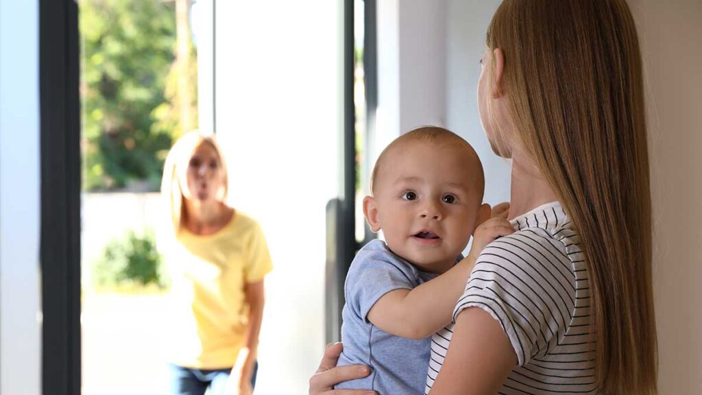 A childminder holding a baby as a woman looks through a door in the background