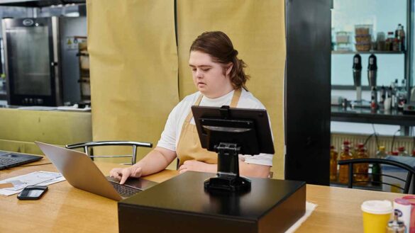 A woman with Down Syndrome working on a laptop beside a till in a cafe
