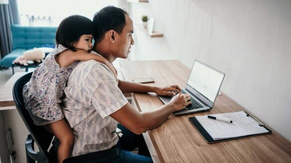 A father working at a desk with a young girl sitting on his back