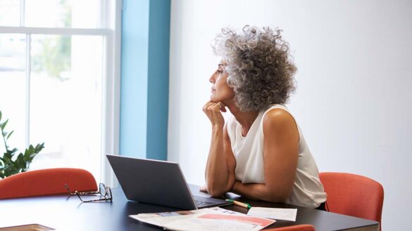 A middle aged woman working in an office, looking out of a window with a laptop on a table