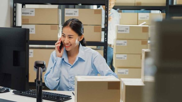 A happy female employee laughing while talking on a phone and sitting at a desk in a warehouse