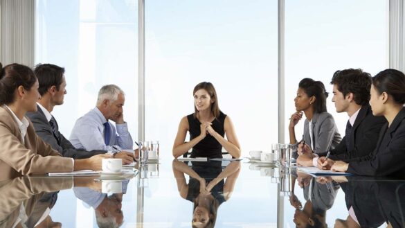 A group of business people around a boardroom table, with a woman at the head of the table
