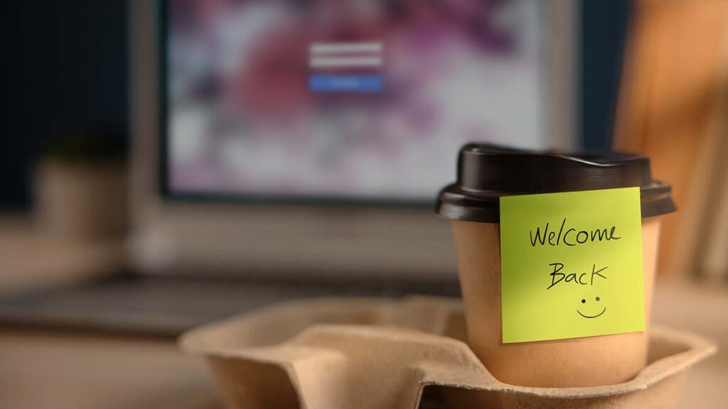 A coffee cup with a note saying 'welcome back :)' stuck to it. It is on a desk with a computer in the background.
