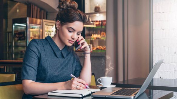 A woman in a cafe. She is writing notes while speaking on a smartphone, with a laptop and coffee on the table in front of her