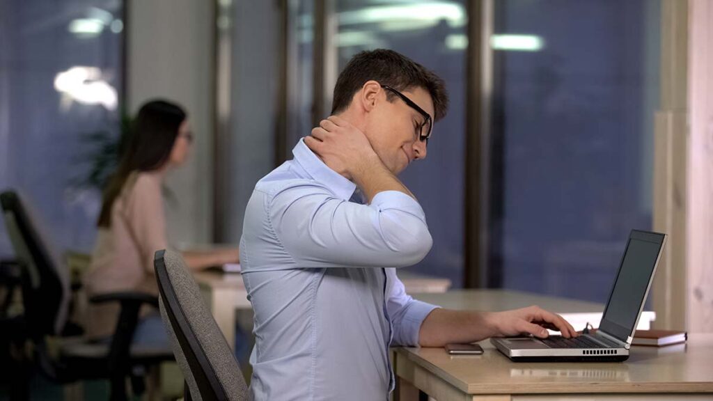 A man rubbing his neck while working on a laptop in an office