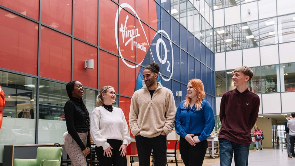 A diverse group of five young people standing in an atrium, all smiling and looking at each other. They are standing in front of a wall with the Virgin Media O2 logo printed on it.