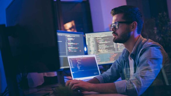 A man surrounded by several computer monitors with code on them.