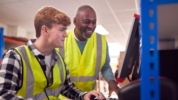 A man working with a younger apprentice on a computer. Both are wearing hi-vis jackets.