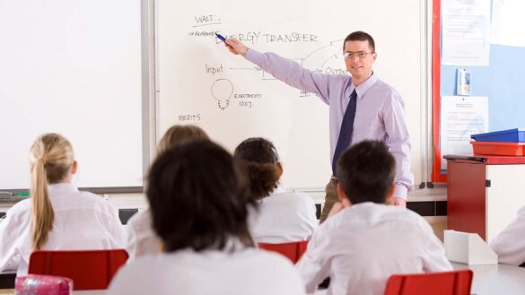 A male school teacher pointing to a whiteboard in front of a classroom of students