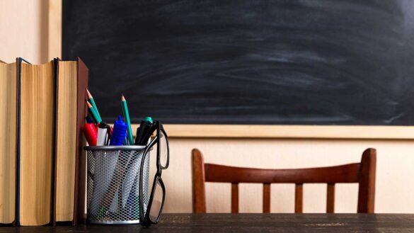 A teachers' desk in a classroom, featuring a pair of glasses, pot full of pens, books and a blackboard in the background