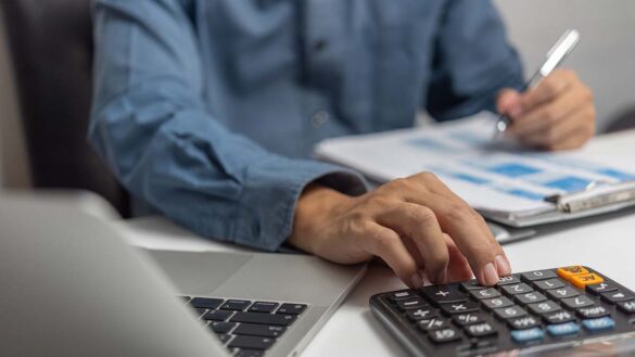 A man using a calculator on a desk with a laptop and paperwork
