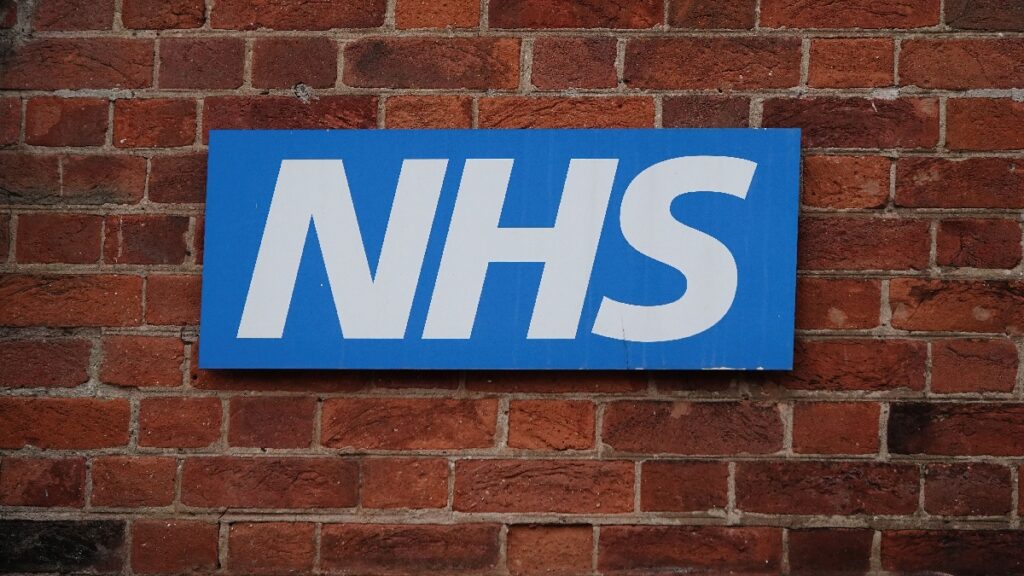 NHS job cuts. Photo shows an NHS sign on a brick wall.
